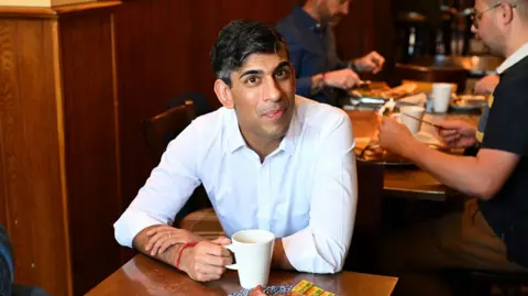 A man with dark hair and wearing a white shirt is sitting at a table holding a white cup.
There are people eating in the background.