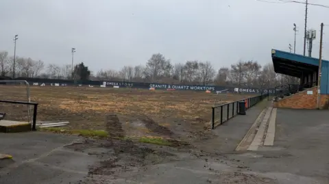 BBC A pitch which is mostly mud, surrounded by advertising boards and lights. A spectator stand on one side of the pitch.