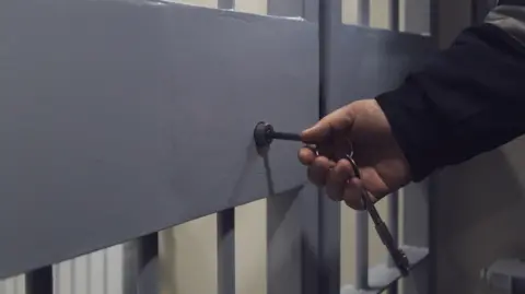 A stock image showing a grey metal barred door with a hand putting a key into a lock on it. The person's arm can be seen and they are wearing a dark long-sleeved jacket.