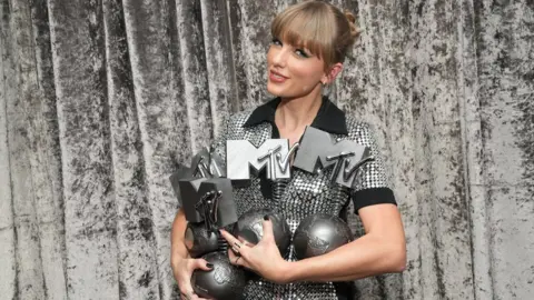 Taylor Swift in a sparkly dress, holding three MTV EMA awards