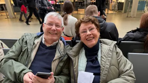 An elderly couple sit at a train station. Both the man and the woman are wearing green coloured jackets and are smiling at the camera. 