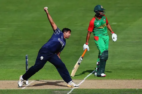 Getty Images Majid Haq of Scotland bowls during the 2015 ICC Cricket World Cup match between Bangladesh and Scotland at Saxton Field on March 5, 2015 in Nelson, New Zealand. He is side on, wearing blue and bowling full pelt at the other wicket from left to right of shot. A Bengladeshi player in green and red is standing behind him at the other wicket.