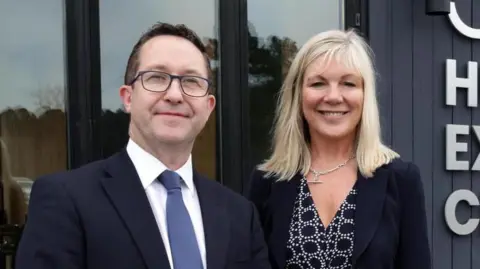 A man and a woman are looking into the camera. The man on the left has glasses and dark hair. He is wearing a dark suit, white shirt and a blue tie.
The woman has shoulder length blonde hair and is smiling. She is wearing a dark patterned dress and dark jacket. She has a silver T-bar chain around her neck