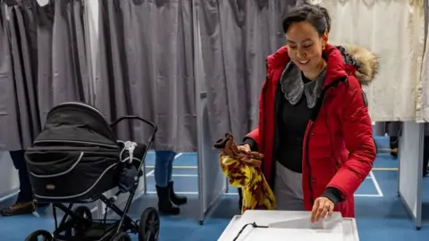 A voter dressed in a red coat casts her ballot in Nuuk while her baby waits near by in a buggy