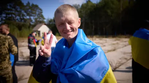 Ukrainian government A Ukrainian servicemen released during a prisoner exchange with Russia is seen squinting and smiling into the camera. He is also wrapped in a Ukrainian flag and holding up two fingers facing forward in a V sign. 