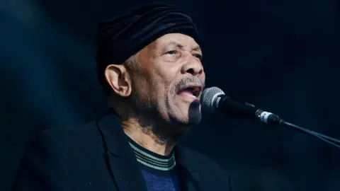 Roy Ayers performing at Glastonbury in 2019. He has a blue top and black jacket on and is singing into the microphone on stage.