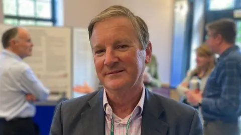 Councillor Nick Botterill looks at the camera - it is a head and shoulder shot while at an event. He wears and grey suit jacket and pink check shirt, and has grey hair.