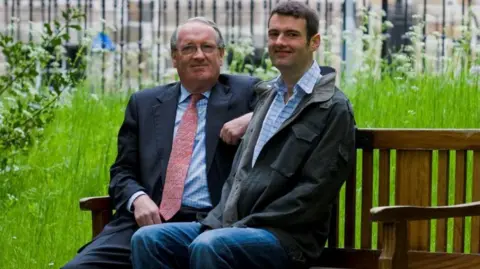 University of Edinburgh Two men, Donald MacDonald and Euan MacDonald sit on a bench. The man on the left has glasses and is wearing a navy suit, blue shirt and a pink tie. The man on the right is wearing jeans, a denim jacket and a blue checked shirt.