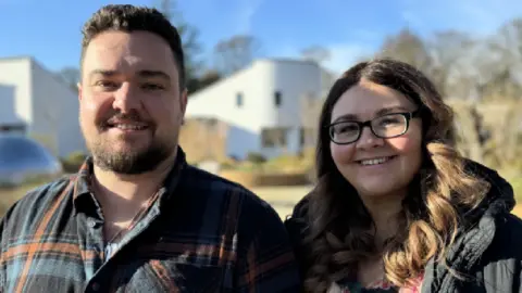 Emily and James Taylor looking at the camera smiling. The picture is taken outside on a sunny day with the hospice in the background. 