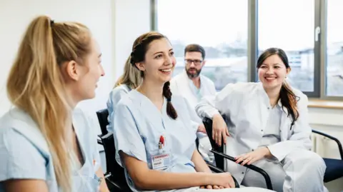 Three women doctors sit on chairs smiling and chatting. 