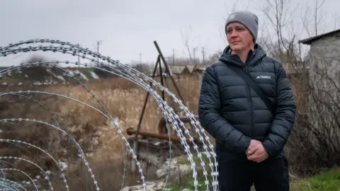 BBC/Matthew Goddard Oleksandr, a man wearing a hat and a dark jacket, stands next to barbed wire.