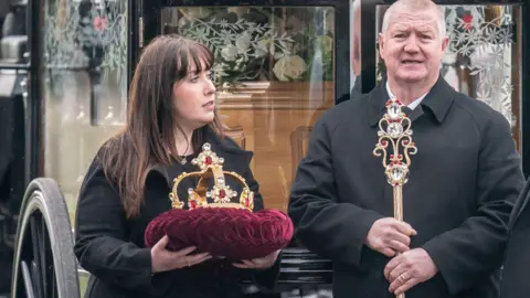 PA Media An ornately designed sceptre and matching golden crown - resting on a purple velvet cushion - which were won on RuPaul's Drag Race UK. They are being carried by a man and a woman into The Vivienne's funeral service.