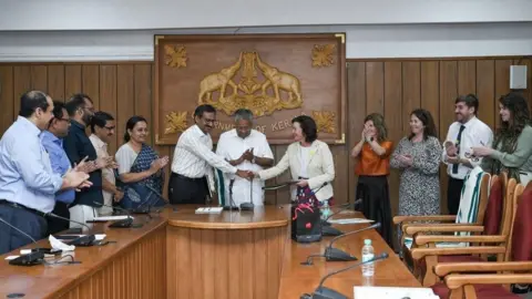 Eluned Morgan, when she was the Health minister,  and a Welsh government delegation meeting meeting there counterparts in Kerala India they are at the had of a push wooden board room with a wood engraved placard on the wall above them. 