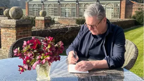 Starmer sitting outdoors writing a letter to his daughter. On the table, there is also a glass vase with pink flowers. Behind him, there is a mowed lawn and a building.