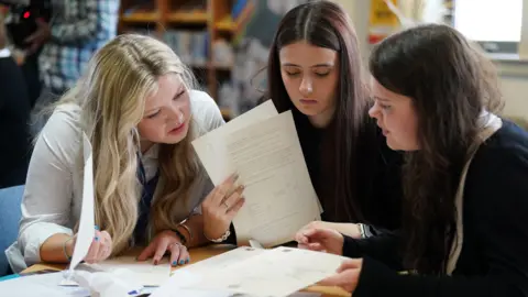 Three female school pupils receiving their exam results. They are sitting at a table looking at their certificates.