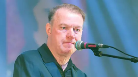 Getty Images Edwyn Collins sitting on stage at a microphone - he is wearing a blue jacket and a green T-shirt and has thinning brown hair