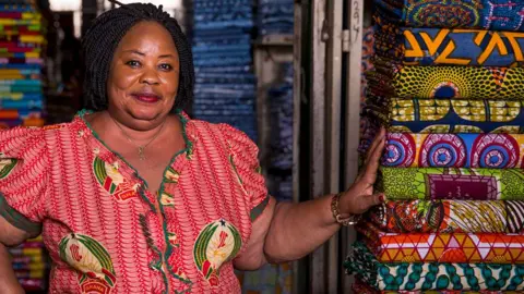 Getty Images A woman in a red print design shirt standing next to wax print rolls of fabric at a stall in a market in Ghana