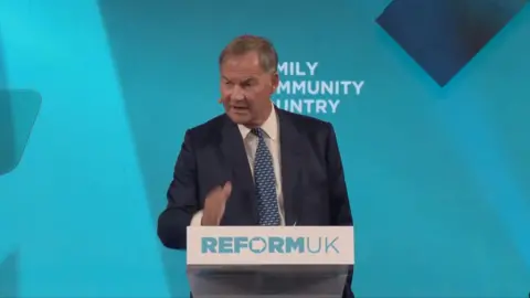 Rupert Lowe speaking behind a lectern at Reform UK event, in front of a bright blue background