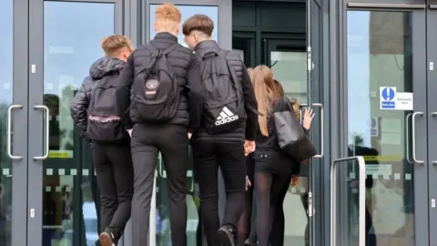 Getty Images A group of three school boys dressed in black and carrying school bags walking through a school door