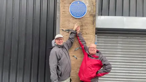 Simon Thake Two men in winter jackets point up to a blue plaque on a brick wall above them 