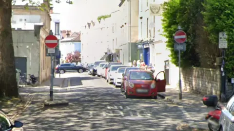 Google Two no entry signs sit on either side of the end of a one-way road. The road has cars parked along one side of it and has one lane for vehicles to drive along.