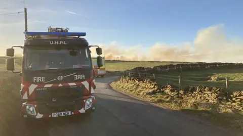 Fire engine on rural road with smoke on moorland in background