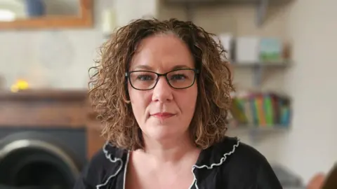 Gemma Whysall is wearing black glasses and has shoulder length curly hair. She is wearing a black blouse with a white trimmed frilly collar. She is looking directly at the camera and there is a book case and a fireplace behind her.