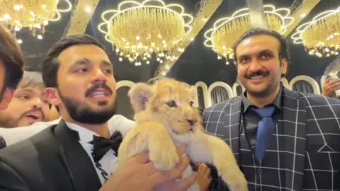 YouTube / Rajab's Family Rajab Butt, in a suit, holding a lion cub. Umar Dolla, in a suit, is stood next to him smiling