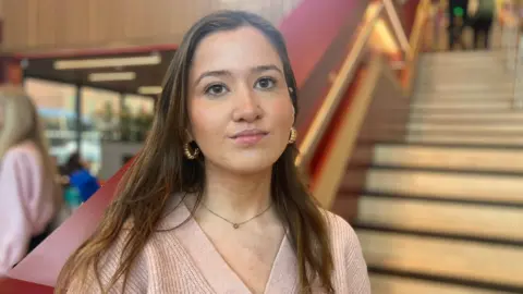 BBC A woman with long brown hair looks into the camera. She is smiling with her mouth closed and is wearing a light pink cardigan and gold hoop earrings. 