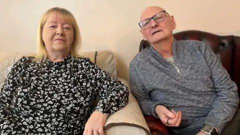 A photo of Florence and Peter sitting a living room. Florence is on a sofa with her arm on the arm rest. Peter is in a leather armchair and is leaning slightly to get in closer to Florence in the photo. He is bald with glasses and grey zipper sweater. She has shoulder-length blonde hair and floral dress.