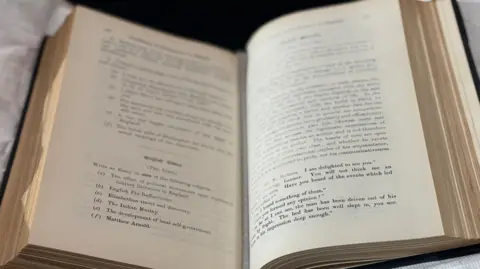 BBC A book is displayed on a protective cushion. It is open, showing some of the questions from the first Cambridge English exam in 1913.