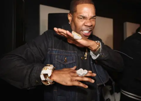 A man, Busta Rhymes, crosses his arms. He wears a denim jackets and gold jewellery.