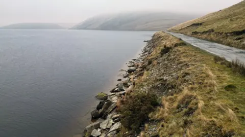 An aerial photograph showing the shore of the reservoir stretching away into the distance. The shore is low and flat, with large stones at the end of the water and a grassy area behind them, leading to an unmade road. 