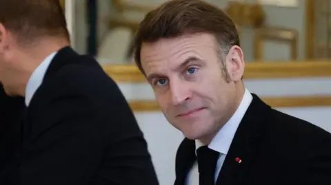 Reuters Emmanuel Macron in a black suit and tie looking off camera with a serious face.