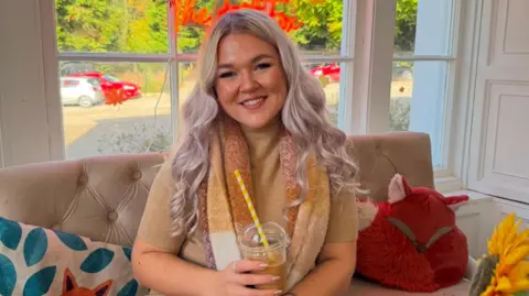 Cara Jackson A woman sitting on a sofa in front of a window smiling at the camera. She is surrounded by colourful pillows. She is wearing a brown short sleeved jumper and a brown, yellow and white scarf. She is holding a plastic cup with a straw sticking out of it.