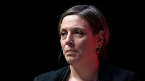 Jess Phillips has short brown hair that is tucked behind her ear. She is wearing a black jacket and is sitting in front of a dark background, She is looking ahead.