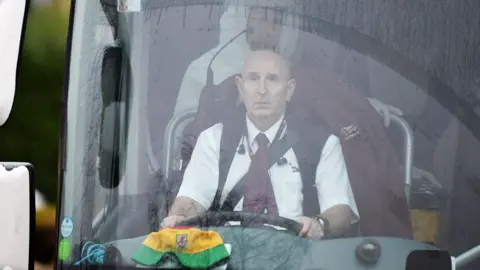 Andy Simonds sitting in the driving seat of the bus he drove, with a bucket Wales cap on the dashboard and people in PPE visible behind him