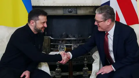 President Zelensky and Sir Keir Starmer shake hands. They are seated in front of their countries flags.