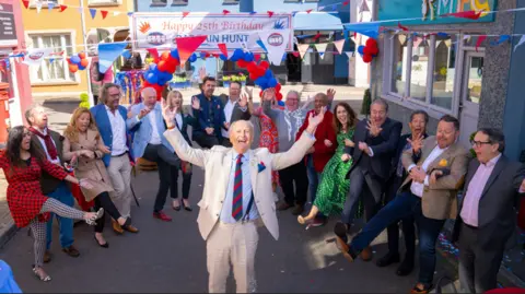 16 experts and presenters from Bargain Hunt form a semi-circle around another of their presenters. The presenter in the middle is a man in his 70s wearing a chalky white coloured suit and light blue shirt. The presenters surrounding him are all doing a kick in the air. They are all standing in a street with blue, white and red bunting under a happy 25th birthday Bargain Hunt banner. 