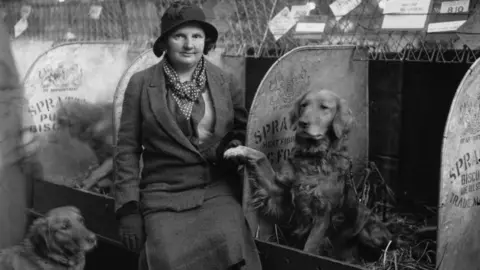 Getty Images A woman is sitting in the dog kennels wearing a dark coloured coat, a spotted scarf and a black hat. Next to her is a golden retriever that is holding its paw in the air. 