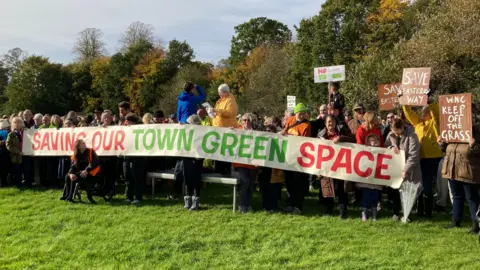 A crowd of people standing behind a banner which says "Saving our town green space". Some are holding placards saying things like "save Eastern Way". One woman is standing on a small platform and reading from a piece of paper whilst wearing a yellow raincoat. They are standing on the grass of a playing field with trees behind.