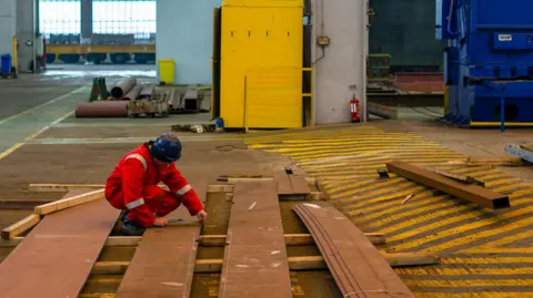 GETTY IMAGES A worker measures pieces of steel for the construction of a barge at the production facility in Harland & Wolff Group Holdings Plc shipyard in Belfast. He's wearing a black hardhat and an orange high-viz clothes. There are pieces of tubes and wood lying about the floor.