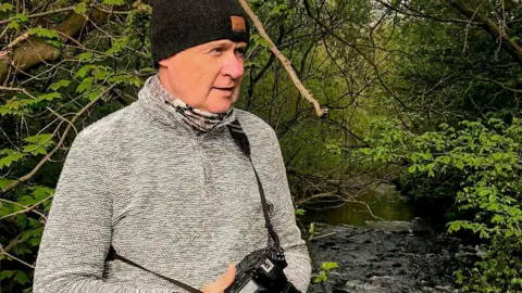 A man standing next to a river surrounded by trees and greenery. He has a camera slung round his body and is wearing a light grey zipper and a black beanie