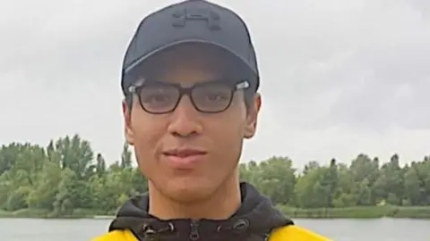 Nicholas Prosper dressed in glasses, a black baseball hat and yellow and black hoodie while standing in front of a lake.