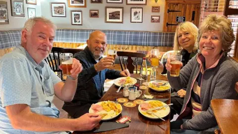 BBC/JULIA LEWIS Four customers, two men and two women are sitting at a table in a pub eating ham, egg and chips.
They have paused to raise their glasses of drinks.