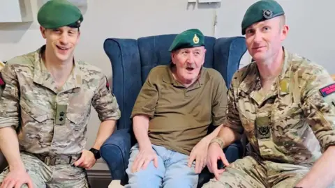 Three Royal Marines. In the middle an older man wearing a green t-shirt and blue jeans. All of them have green beret's on. 