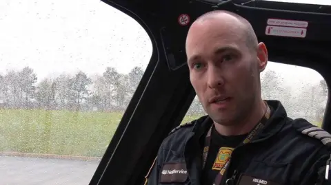 A man in black pilot overalls sits inside a helicopter cockpit with rain streaks on the windows.