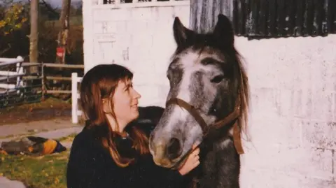 Ginny gazing up at a grey and white horse in the yard. Ginny has long brown hair and a fringe and is wearing a dark jumper. 