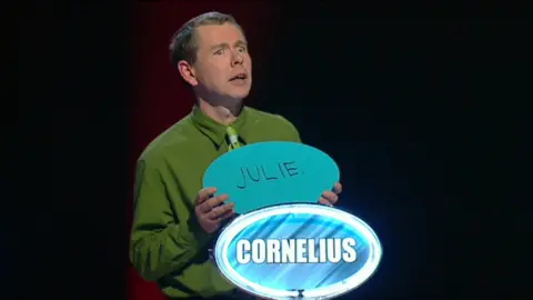 The Weakest Link/BBC Cornelius O'Donovon wearing a green shirt and tie appearing on the Weakest Link with Anne Robinson. There is a sign in blue and white with his name on it and he is holding a sign with the name Julie on it.