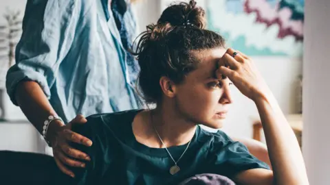 A sad-looking woman rests her forehead on her hand, while someone in the background puts a hand on their shoulder. 